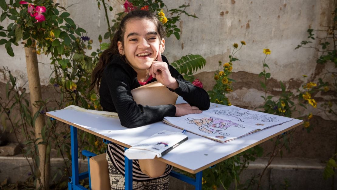 An Afghan teenage girl stands in a special standing frame with a desk. She is smiling. A coloring book is open on the desk.
