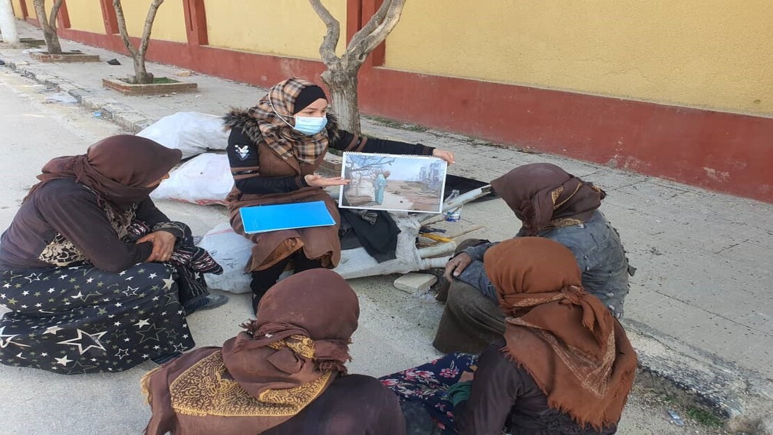 Group of four women, all wearing head coverings, sitting on the street / sidewalk. In the middle, a woman holds up an image as part of a risk education session