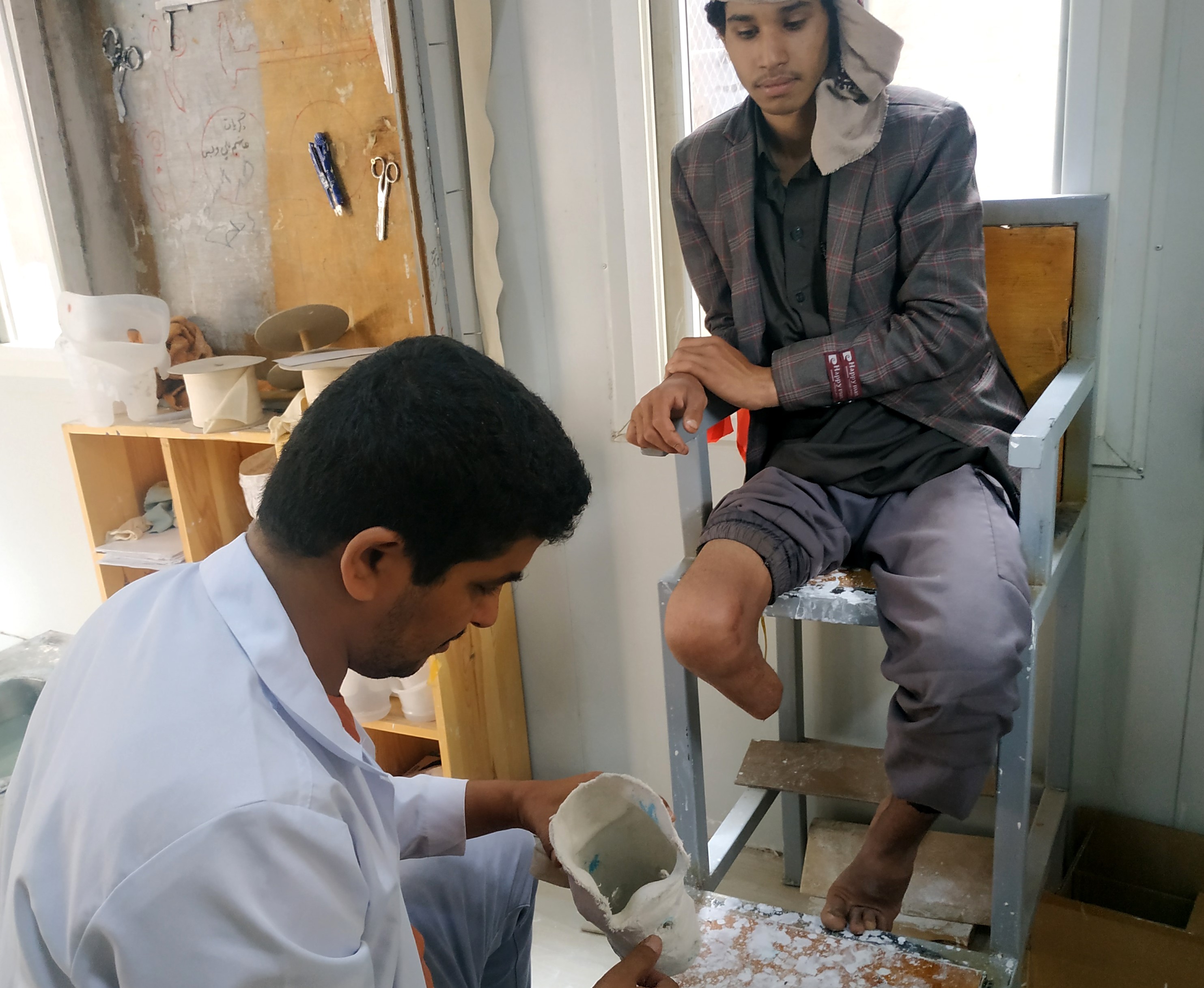 Teenage boy with amputated leg sitting in a chair. Prosthetic technician kneels in front of the boy, holding a prosthetic leg 