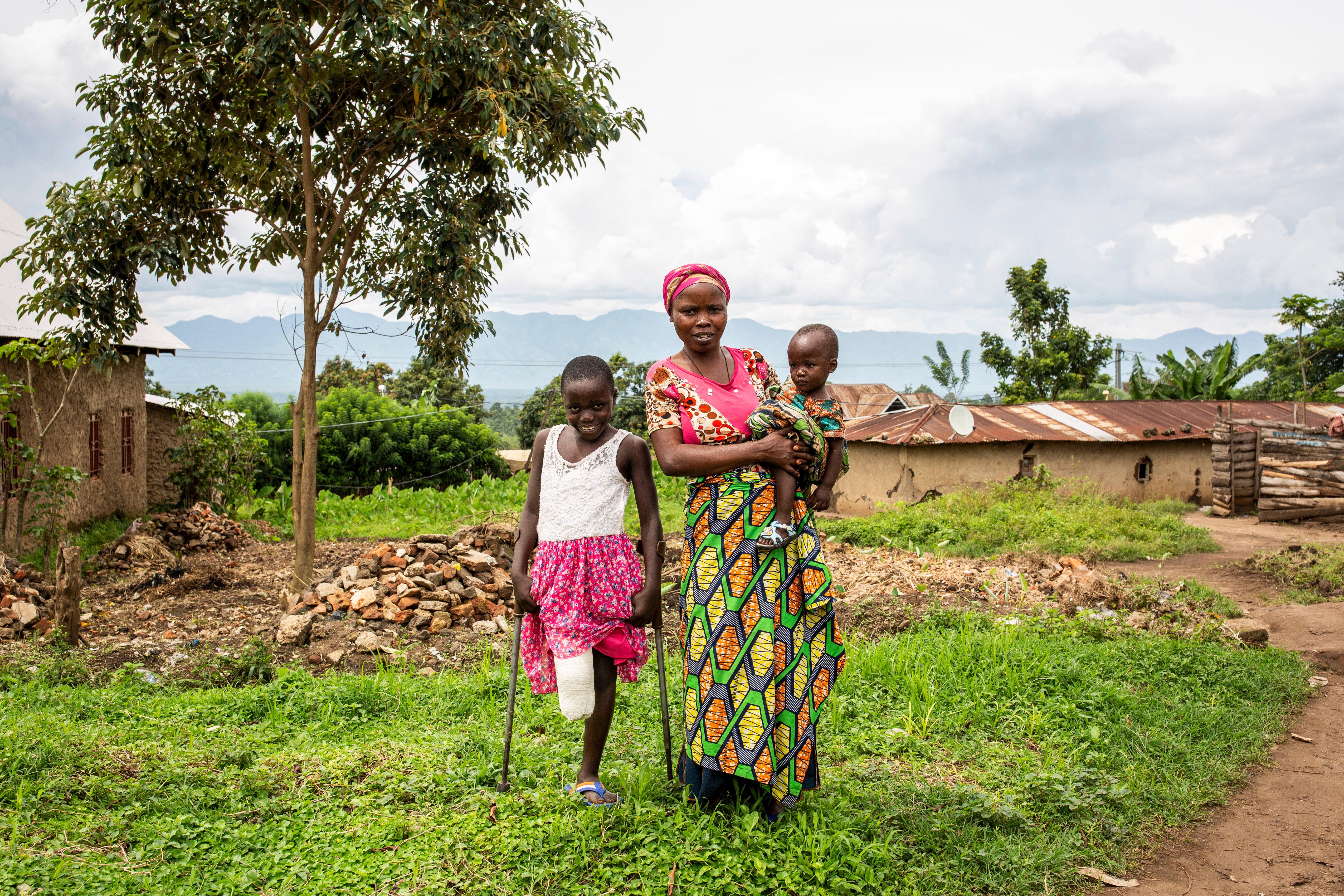 A woman with her two children. She holds a baby in her arms and is next to a young girl on the left who smiles as she uses crutches to support her one leg. 