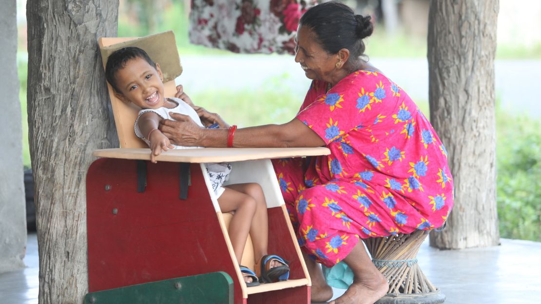A young Nepali girl sits in a specially adapted chair. Her grandmother, wearing a pink floral dress, sits on a stool beside her. They are both smiling.