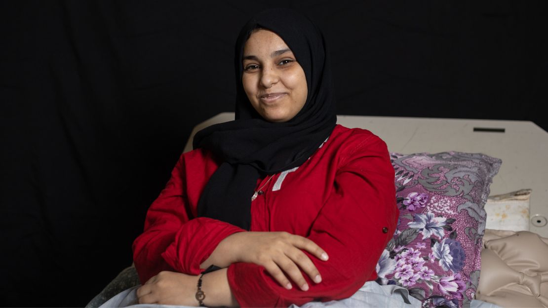 A woman wearing a red blouse and black headscarf sits in a bed with a slight smile across her face.