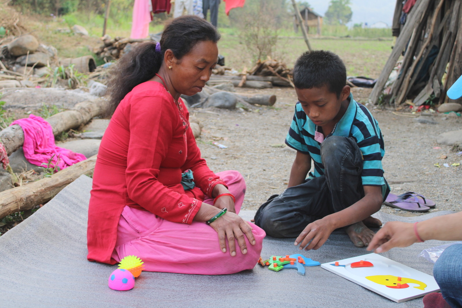 A woman and young boy sit on a mat outside, playing with educational toys