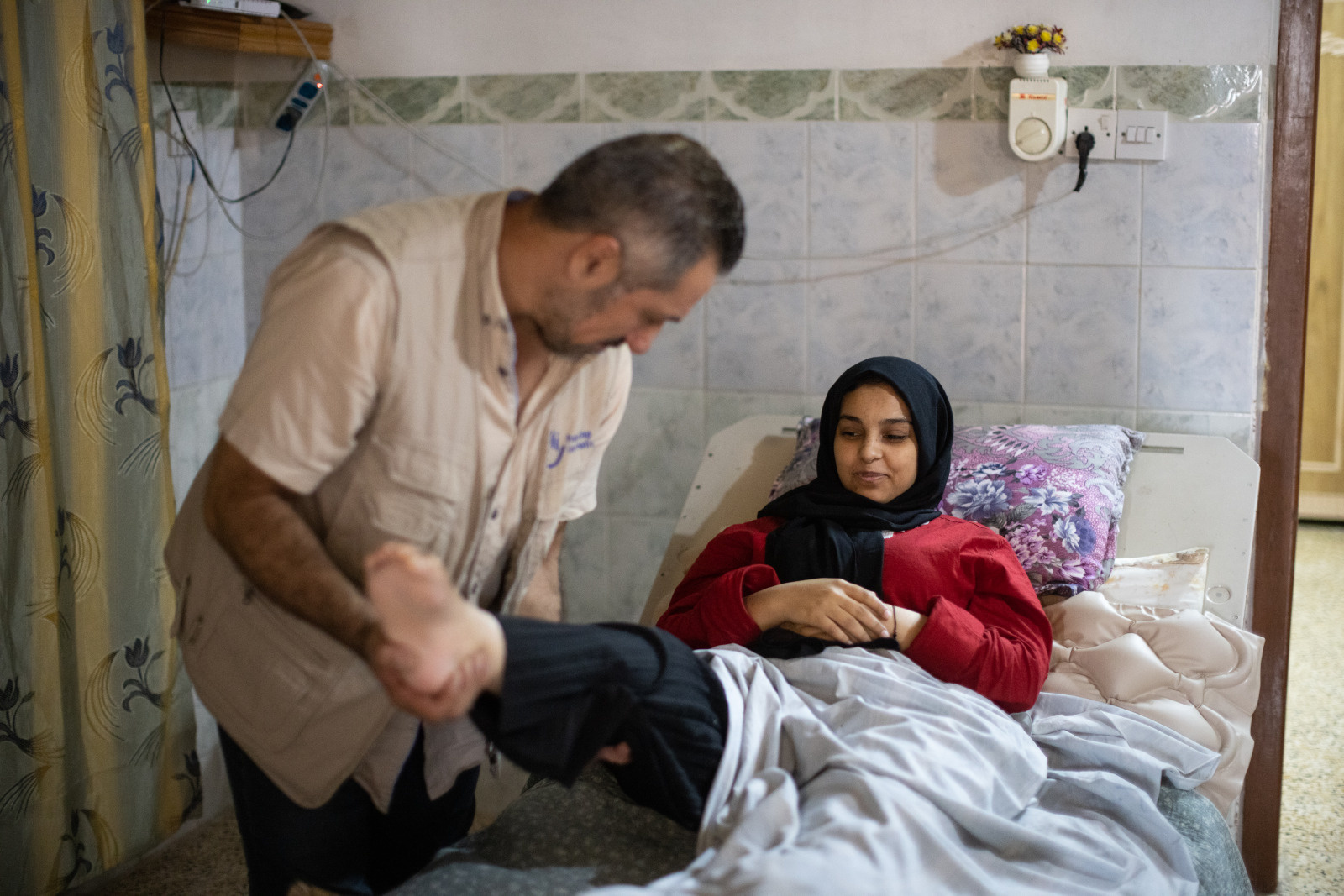 An HI physical therapist works with a patient who is lying in bed. He lifts her leg during exercises.