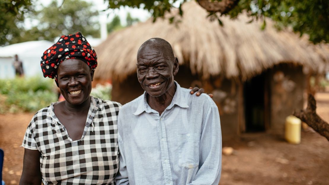 A black woman wearing a black and white checkered shirt and head wrap rests her hand on her husband's shoulder. He's wearing a blue collared shirt. They are both smiling.