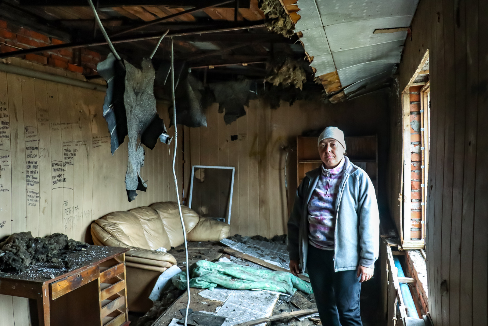 A woman wearing a coat and hat stands in a room in a destroyed building. The ceiling is falling in.