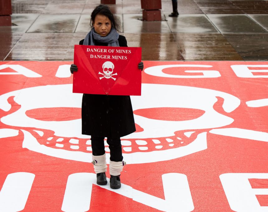 A Cambodian woman wearing a coat and boots holds a red sign that reads Danger Mines with a skull