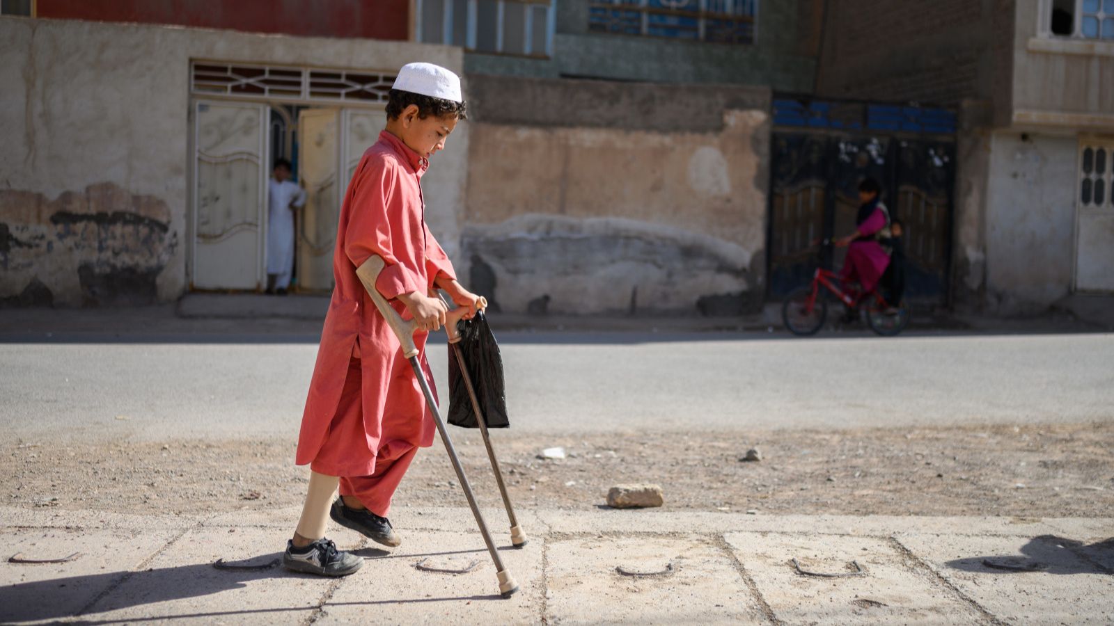A young Afghan boy wearing a coral outfit and an artificial limb walks down a sidewalk with elbow crutches