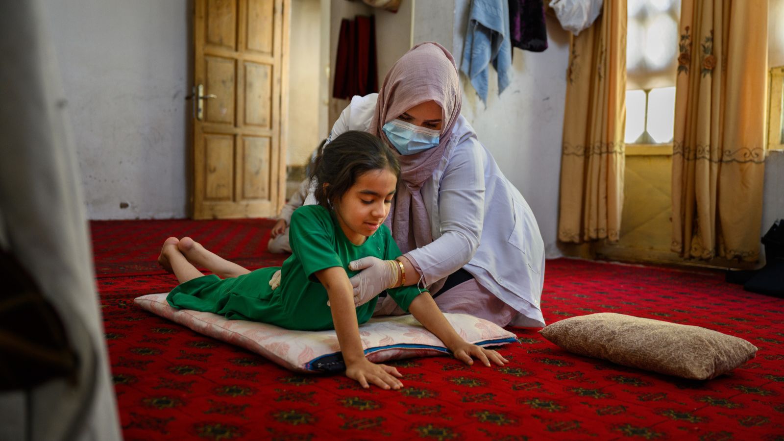 An Afghan girl wearing a green outfit lies on her stomach on a cushion pushing her upper body off the floor. A rehabilitation specialist wearing a head scarf and mask supports her chest.