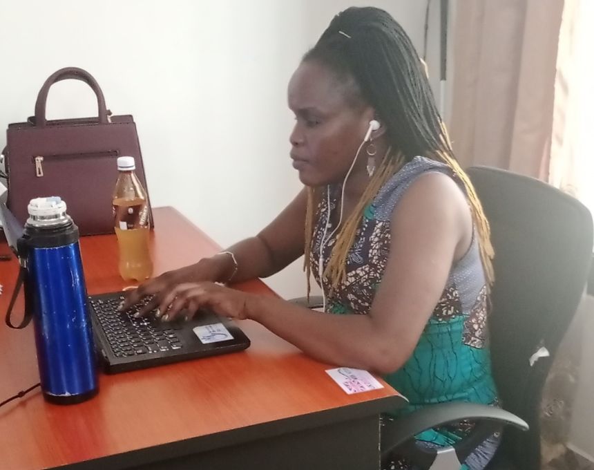 A Black woman sits at a computer desk working on a laptop