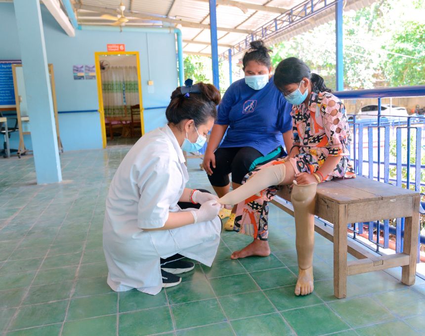A Cambodian girl sits on a bench beside her mother while a technician fits her for an artificial limb