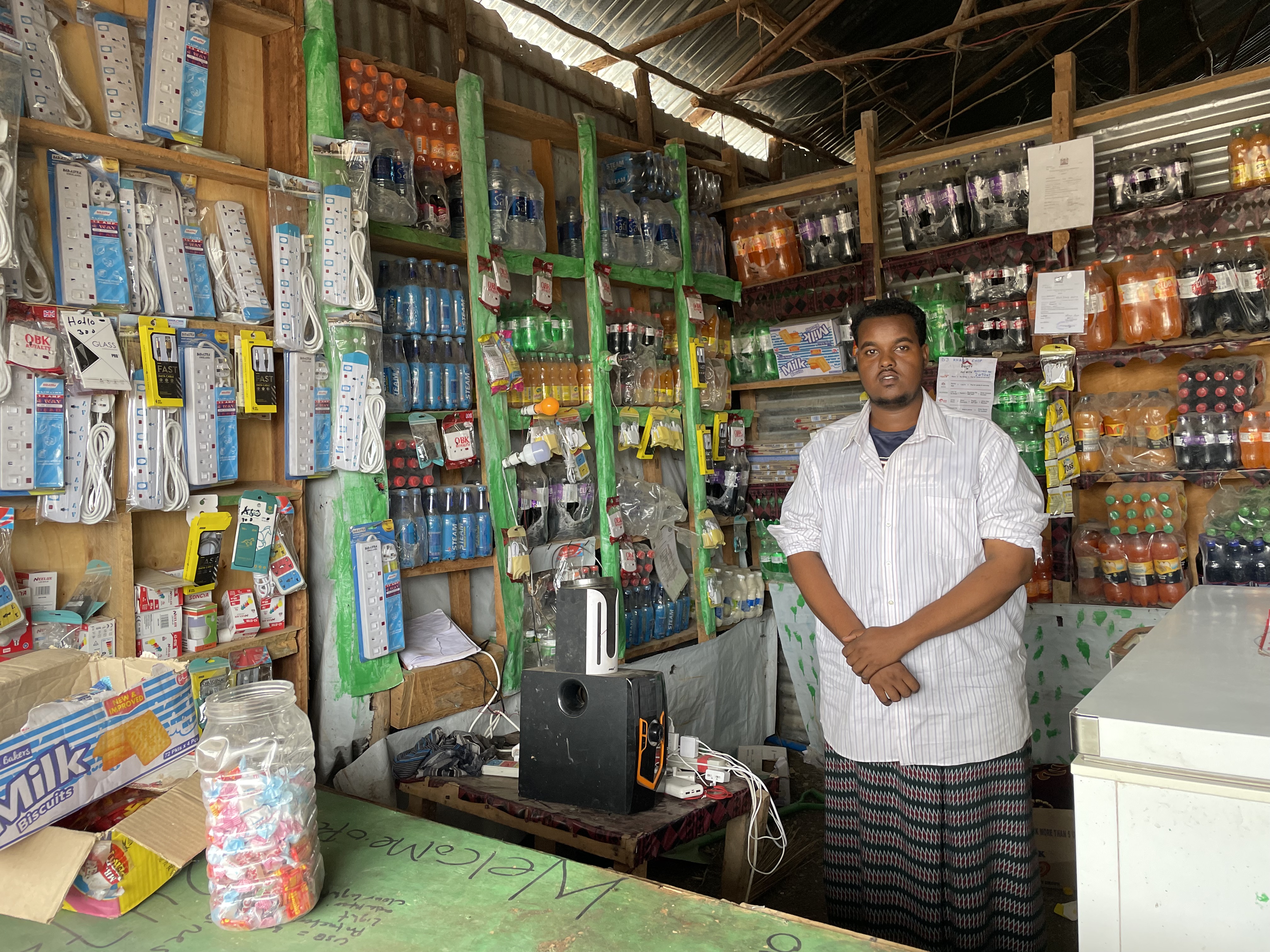 A man wearing a white button-up stands in a shop with phone accessories and drinks on the shelves