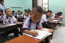 Young boy sitting at a desk in a classroom with children in uniforms. using a pencil in a notebook Nepal