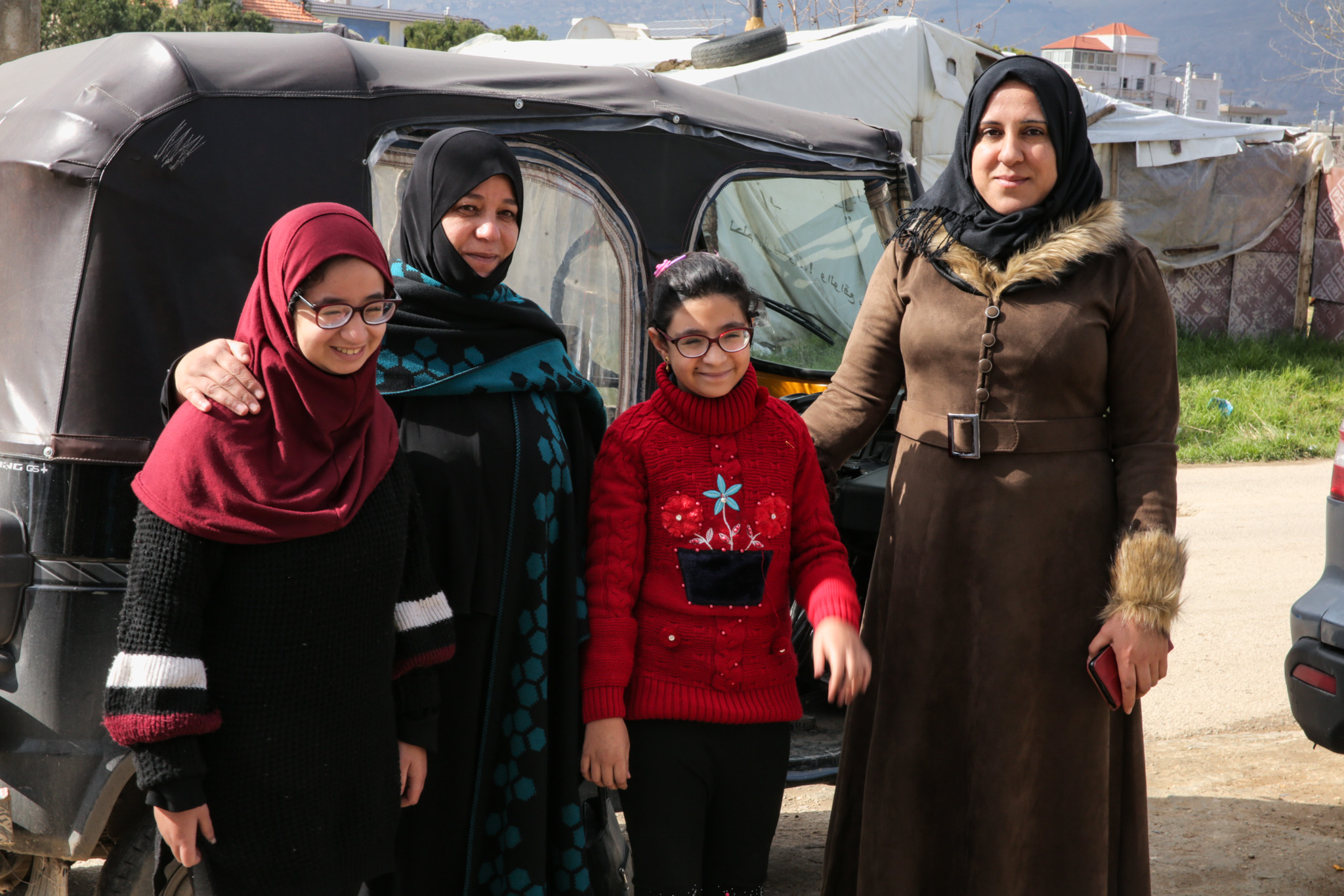 Group of four people smiling, standing in front of tents. From left to right: young girl wearing red head covering and glasses; a woman wearing a black head scarf; a young girl wearing a red sweater and glasses; and a woman wearing a black head scarf and long brown coat.