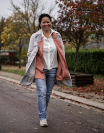 A woman smiling walking down a street, wearing a grey trench coast, pink vest, white button down, blue jeans and white sneakers. 