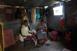 Atilio and his wife in their dimly-lit home, an informal settlement with one window in Maicao, Colombia, sitting down surrounded by houseware and clothing. 