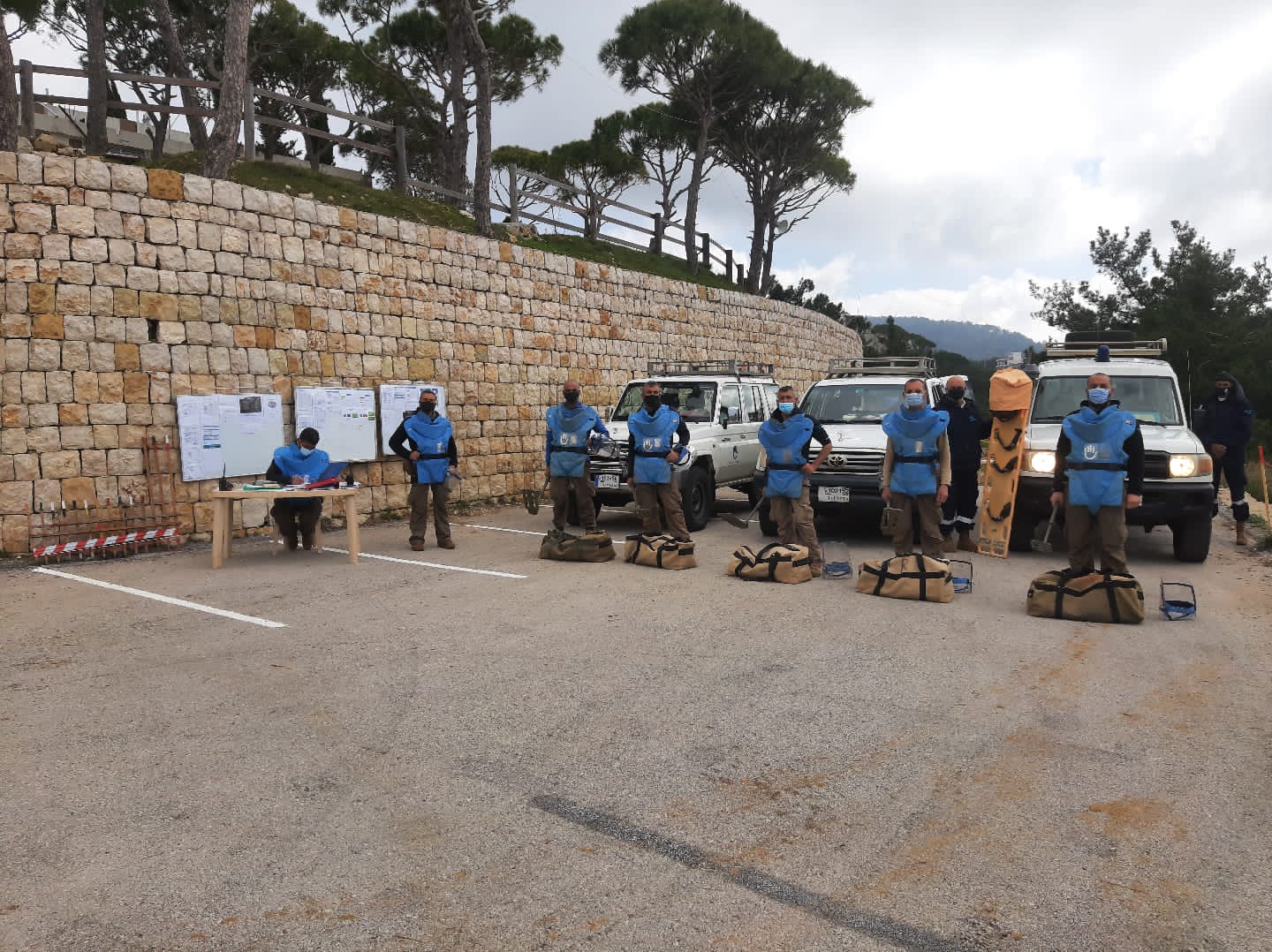 7 deminers wearing blue protective gear, standing in a parking lot in front of 3 vans, next to 3 white boards