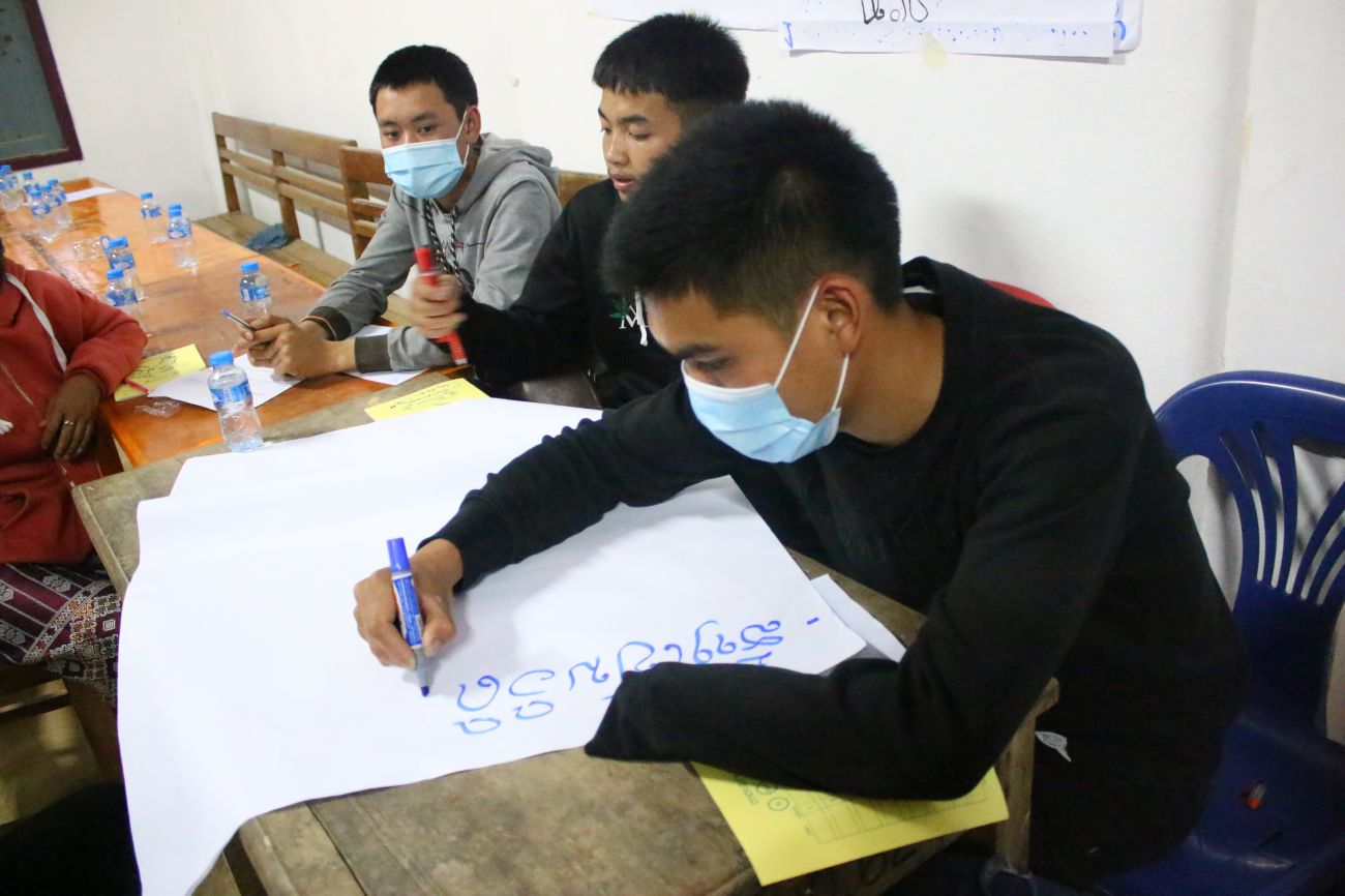 Joc wearing a mask at school writing on a white poster with a market, sitting next to two of his classmates.