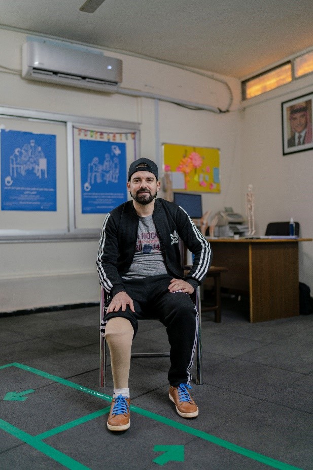 Man wearing baseball hat and track suit sits in a chair placing one hand on his prosthetic leg as he smiles into camera