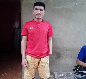 Portrait of Joc, a young man with one arm, looking into camera. Wearing red shirt and khaki shorts against a grey wall background