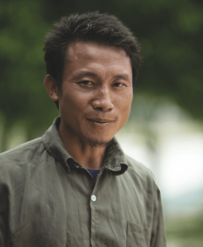 Portrait of Chue Por, a middle aged man, wearing a khaki green button down, smiling in front of a green nature backdrop