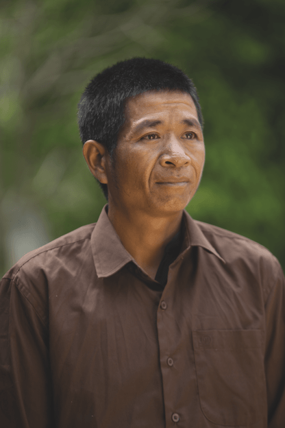 Portrait of Mr. Khamphong, a middle aged man wearing a brown buttown-down, looking off into the distance in front of a green nature background