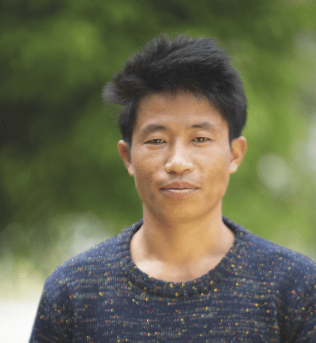 Portrait of Tok, a young man wearing a dark grey/blue sweater, smiling in front of a green nature background.