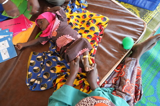 Massalouka, a young girl wearing a pink head covering, lays down on her back during a stimulation therapy session. © J. Labeur / HI