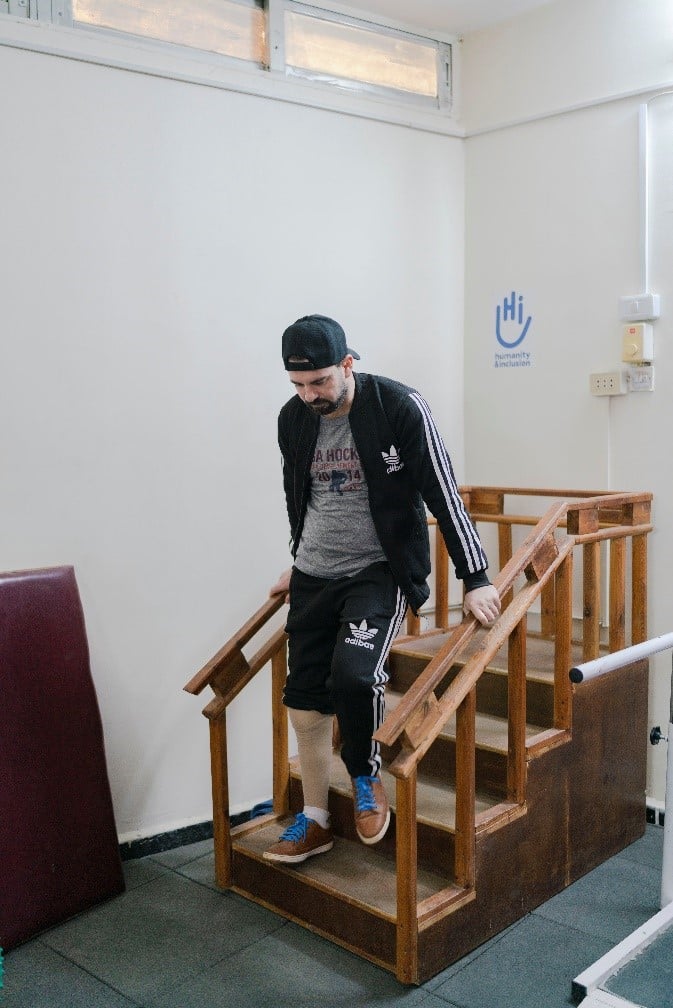 Man wearing baseball hat and track suit walks down model stairs, holding on the railings, to support himself and his prosthetic leg