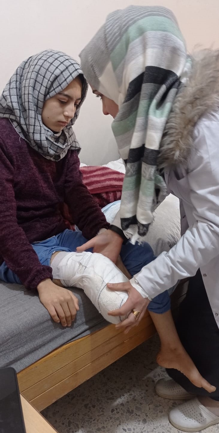 A teenager who has lost her right leg sits on a medical bench, her wound in a cast, being assessed by physiotherapist. Both are female and wearing head scarves. 