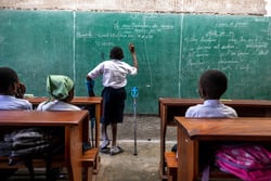 Raphaël writes on a green chalk board during a math lesson in Selembao inclusive school. He stands beside his walking devices. © T. Freteur / HI