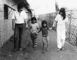 Black and white photo of 4 people holding hands (Jean Baptiste Richardier, Mom Sok, Émilie Vath and Marie Richardier, in 1982). The adults stand on the outsides, the man on the far left, and the woman on the far right. In the middle are two young children. The girl on the inner left has a device on her left leg.