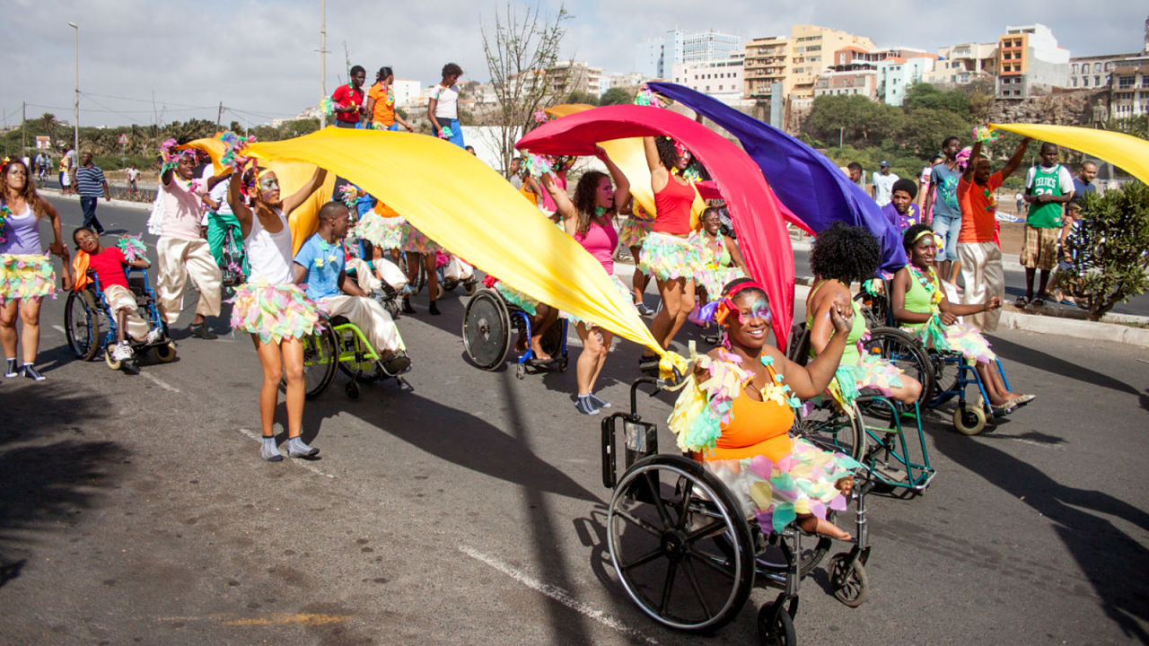 A parade. Group of persons with disabilities, many in wheelchairs, all wearing bright colorful costumes and face paint. Some are waving long pieces of fabric in the air. 