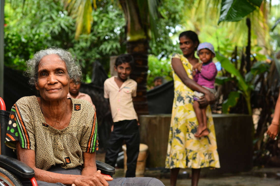 A woman in a wheelchair in the foreground with the rest of her family, including a baby, in the background under a tree.