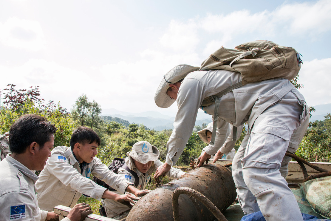 Deminers lift a huge shell together to place it in a vehicle.