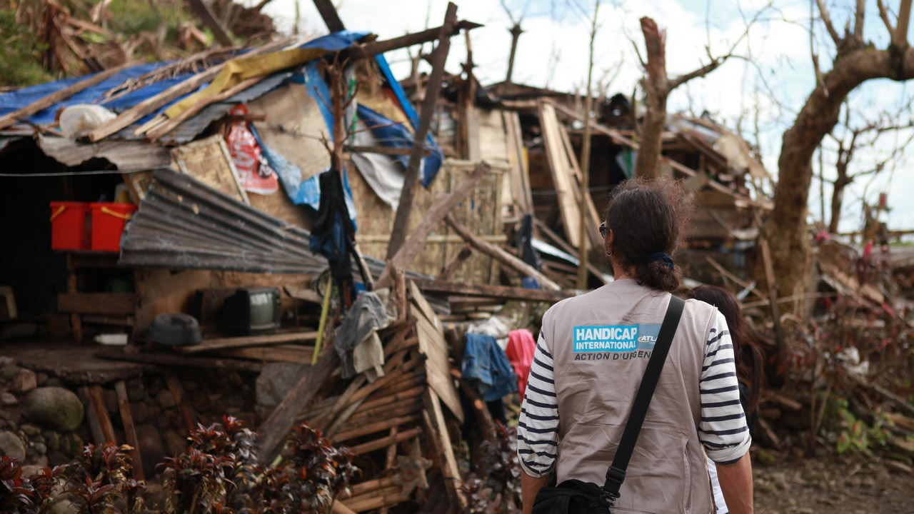 A woman wearing a khaki HI vest amid destroyed buildings in Leyte province following typhoon Haiyan, Humanity & Inclusion Philippines.