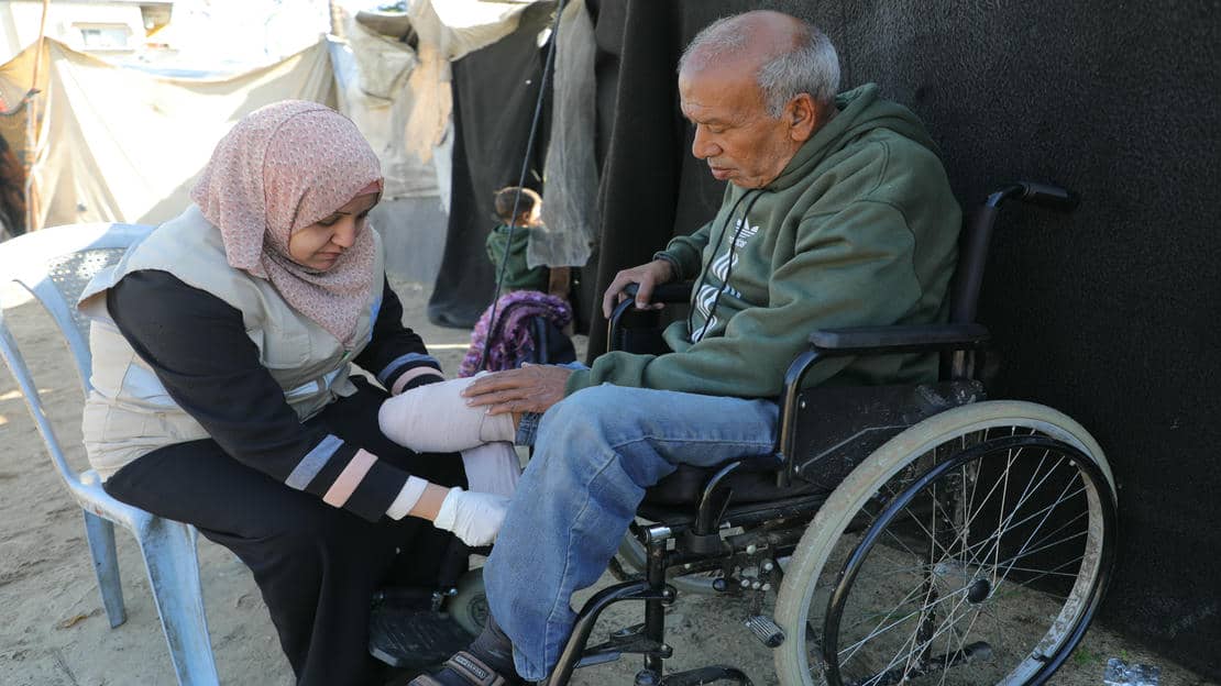 Fayez is sitting in a wheelchair. A woman sitting opposite him on a chair is bandaging his leg.