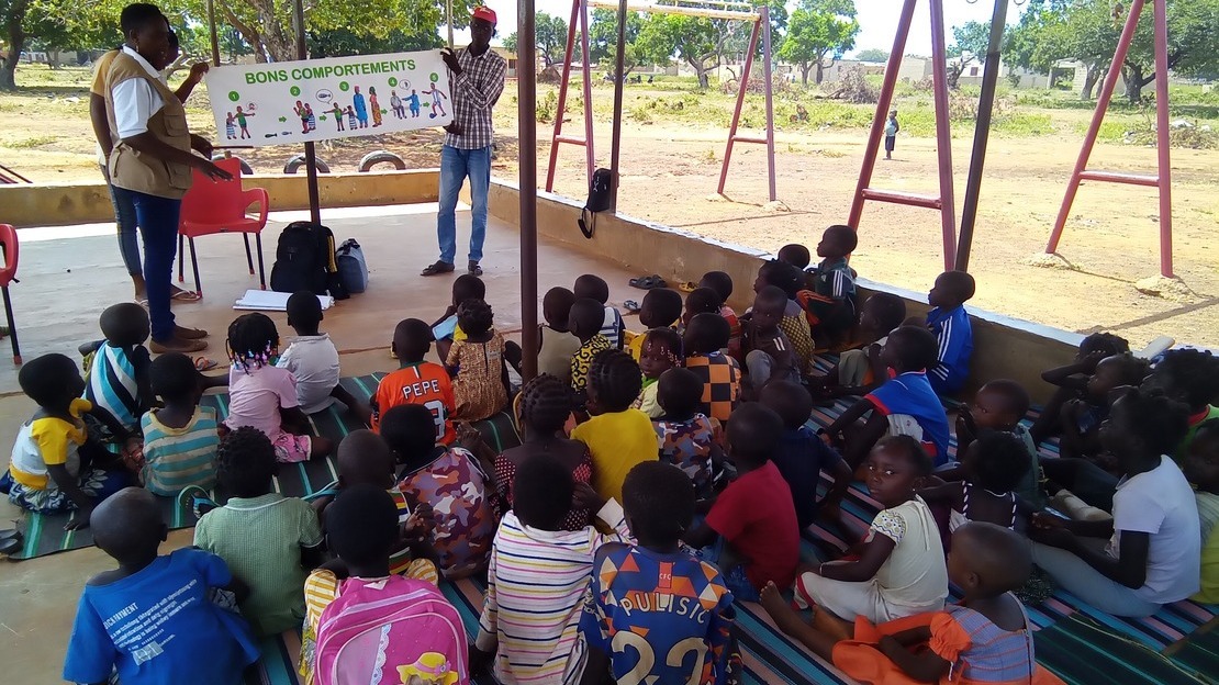 A group of children seated with their backs to the camera look at three adults who show them a banner with diagrams, entitled 