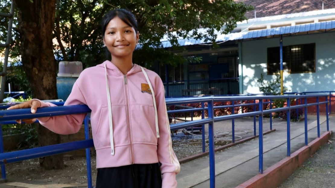 Portrait of Srey Neang leaning against a physical therapy exercise bar in the courtyard of the rehabilitation centre. Behind her, a large tree and an exercise ramp can be seen.