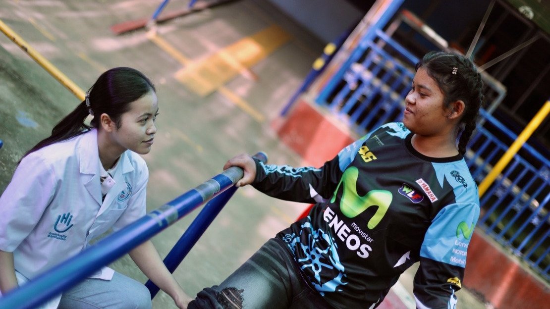 A young girl stands between two physical therapy exercise bars and looks down at a lady crouching beside her wearing a HI-branded smock.