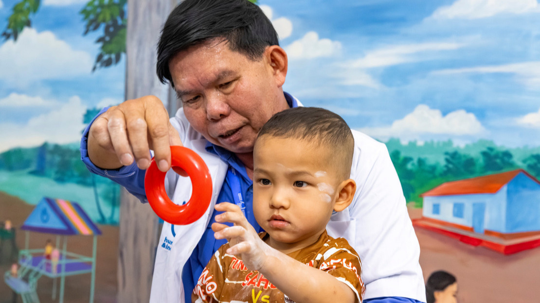 A close-up portrait of a man in a white lab coat holding a young boy on his lap. The boy is reaching out to grab a small plastic ring that the man is holding out in front of him.
