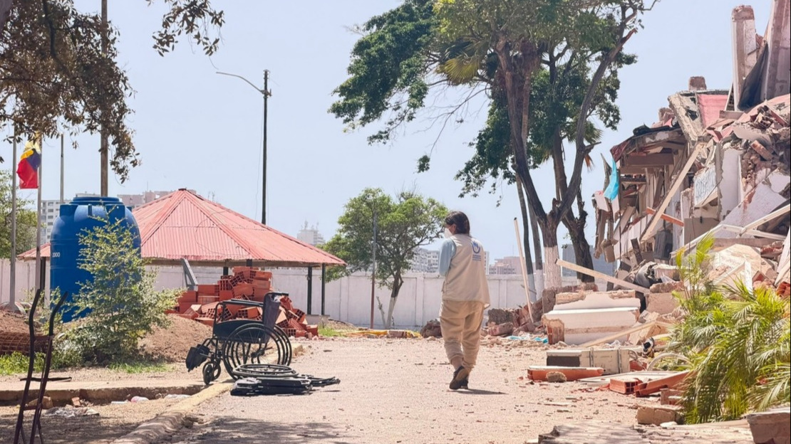 A woman stands with her back to the camera on a dirt road surrounded by building rubble. She is looking to her left at two abandoned wheelchairs.