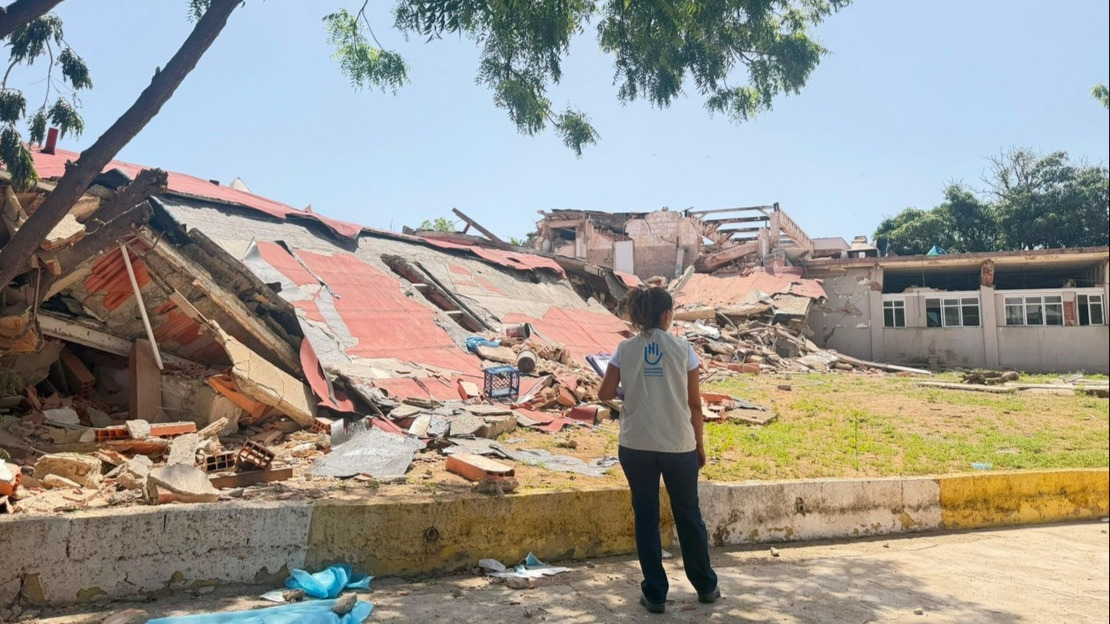 A woman is standing with her back to the camera in front of a completely destroyed building.