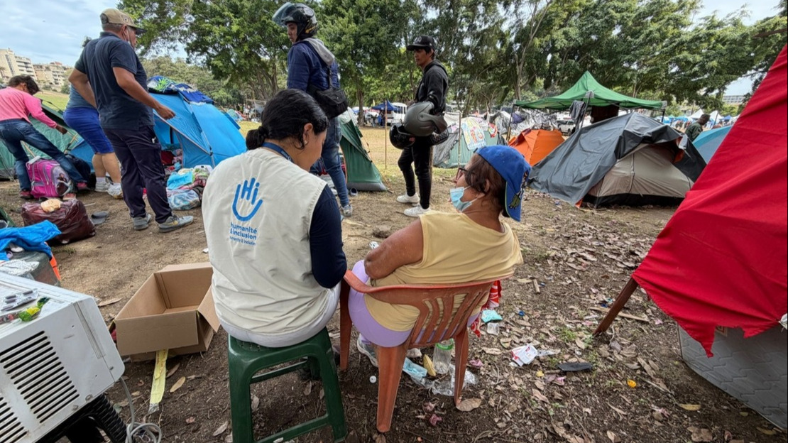 Two women are sitting with their backs to the camera on chairs; one is wearing an HI jacket. Around them are tents, cardboard boxes, scattered belongings, and people chatting.