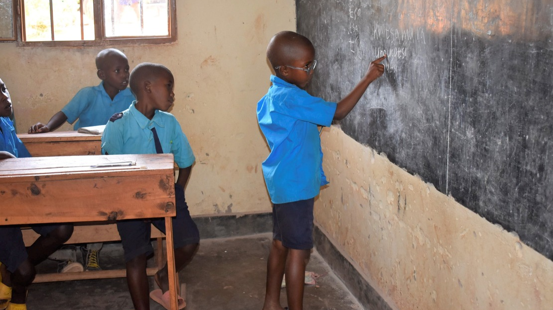 Egide stands in front of a blackboard in a classroom. With his finger, he points to the text written on the blackboard. Two other students are sitting to his left, watching.