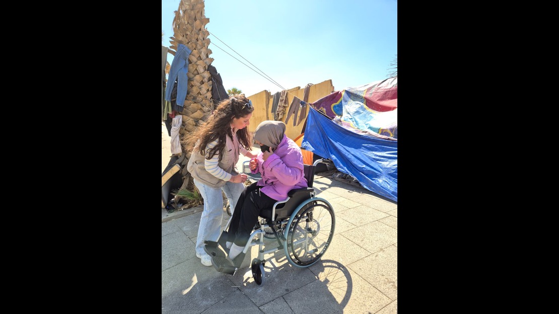 A person is sitting in a wheelchair outdoors, wearing a purple jacket and a scarf on their head. The wheelchair is on a paved surface. A person standing is leaning toward the person in the wheelchair and holding their elbow. In the background, there is a blue tent, clothes hanging on lines, and a palm tree, all under a clear sky.