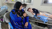 A little boy with bandaged legs is lying on a hospital bed. His grandmother is sitting next to him. The little boy is suffering from multiple fractures and contusions.  