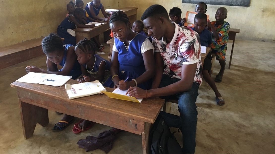 Teenage girl looking to front of classroom. Seated at a desk next to a itinerant teacher that is looking down and pointing to her notebook. In the background are other schoolchildren seated at desks in rows.