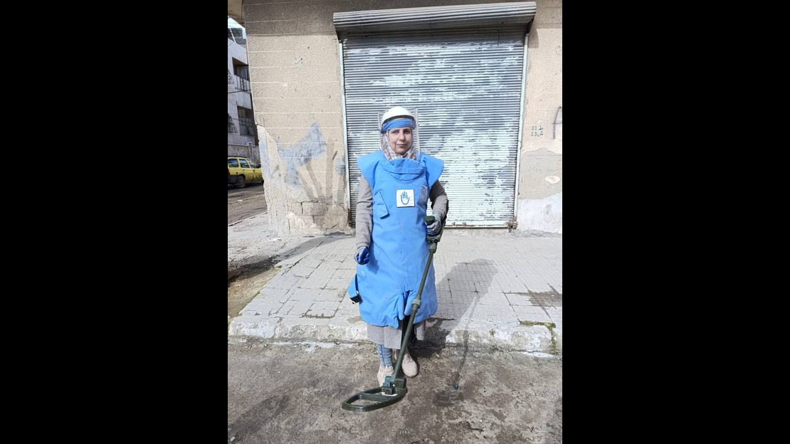 A woman is standing on a sidewalk in front of a closed metal roll-up door. She is wearing a thick blue protective vest and a clear face shield. She is holding a metal detector pointed toward the ground. The building behind her is dilapidated, with partially damaged walls. A yellow car is visible in the background on the left side of the street.