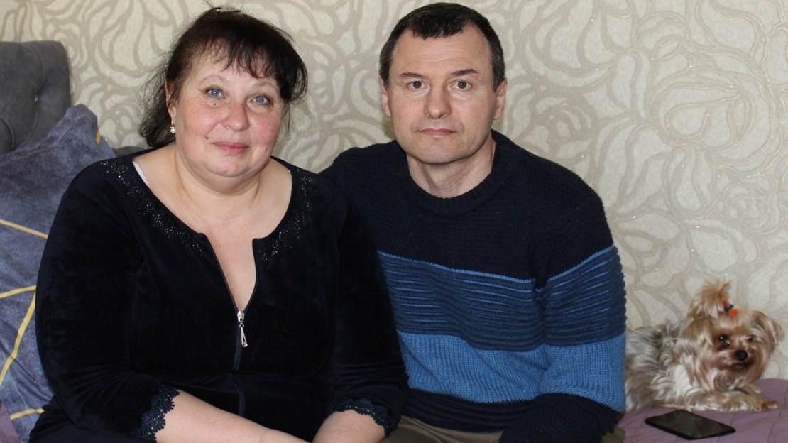 A man and a woman are sitting side by side on a sofa in an indoor setting. Both are facing the camera. The woman is wearing a dark top with a front closure, and the man is wearing a long-sleeved sweater with contrasting stripes. The sofa is covered with cushions, some of which feature various patterns or textures. To the right of the image, a small long-haired dog is lying on a surface near the sofa. Next to the dog, a cell phone lies flat on the floor. In the background, a wall covered in wavy-patterned wallpaper spans the entire width of the image. The lighting is indoor and even, highlighting the faces and objects visible in the foreground.